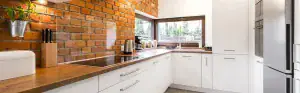 Modern kitchen with white cabinets, wooden countertops, built-in appliances, and a large window. A brick accent wall adds warmth, while kitchen tools and jars decorate the counter. Natural light highlights the space—ideal for easy refrigerator repair access.