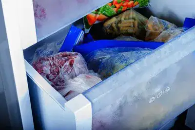 Freezer-Repair An open freezer drawer filled with various frozen foods, including bags of vegetables and other items in plastic packaging, with frost visible on the surfaces.