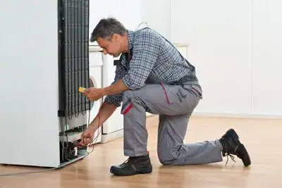 Refrigerator-Repair A repairman in work clothes kneels on the floor, fixing the back of a refrigerator with a screwdriver in a bright, clean kitchen.